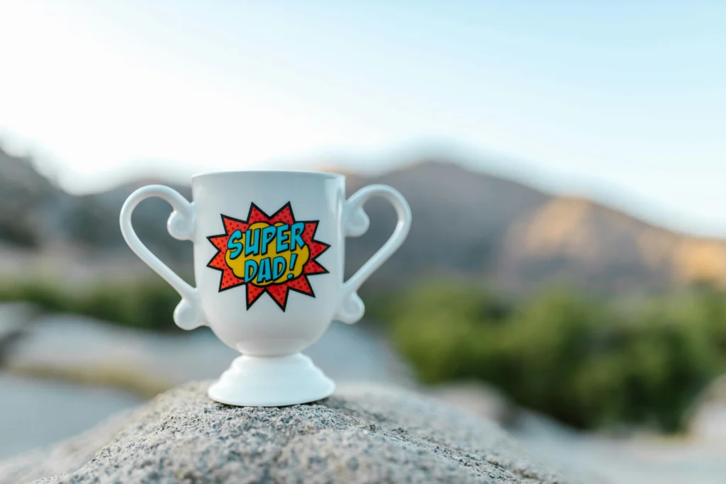 Image of a Trophy-Shaped Mug that Reads Super Dad on a rock with mountains in the background. Image by the RDNE Project via Pexels.com.